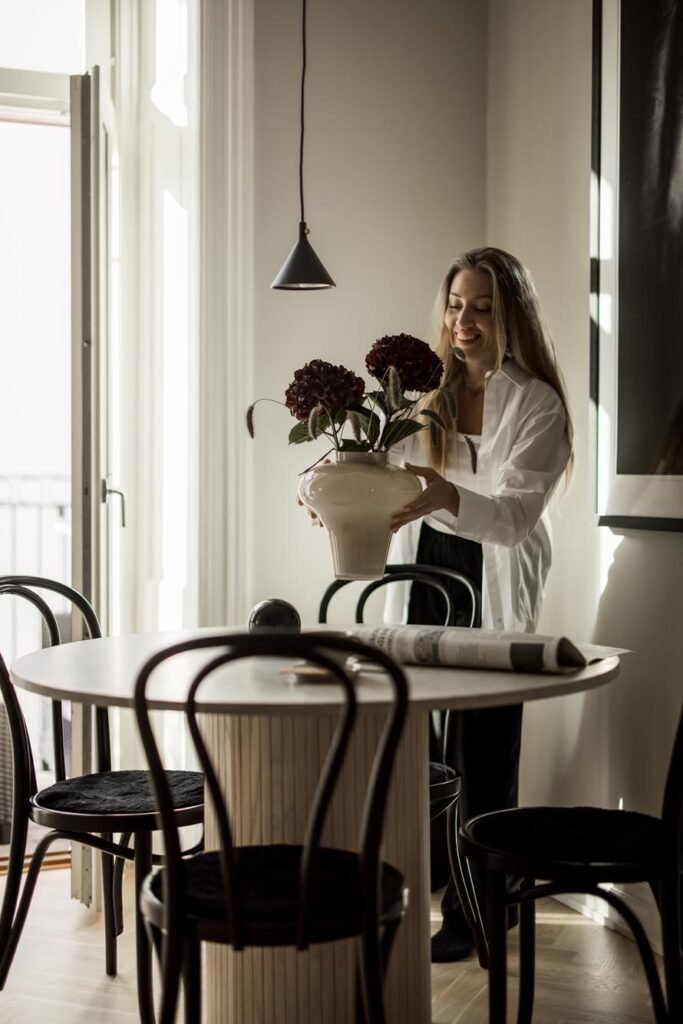 Stiligt - En kvinna ställer en vas med mörka blommor på ett runt matbord i ett modernt, ljust rum - Köket som hjärtat i hemmet - omgivet av svarta stolar och en tidning. Köket som hjärtat i hemmet – Mer än bara en plats för mat: Skapa gemenskap och värme