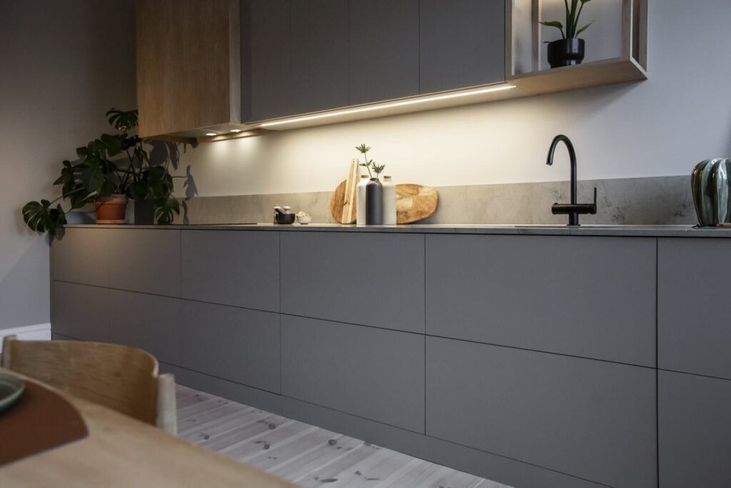 Stiligt - Stiligt och modernt, detta kök har mattgrå skåp, belysning under skåpet, en elegant svart kran och livfulla växter som pryder bänken. Det ljusa trägolvet förstärker den chica atmosfären i förgrunden.