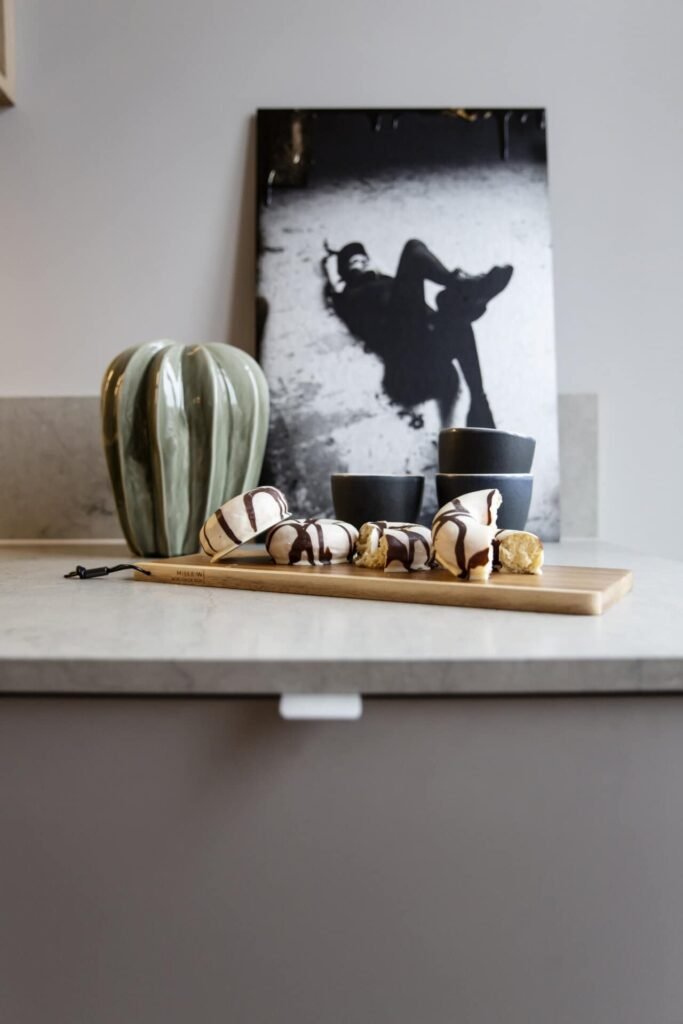 Stiligt - En träskiva med stiligt krämfyllda bakverk översållade med choklad sitter på en bänkskiva bredvid två svarta koppar, en grön vas och ett monokromt konstverk i bakgrunden.