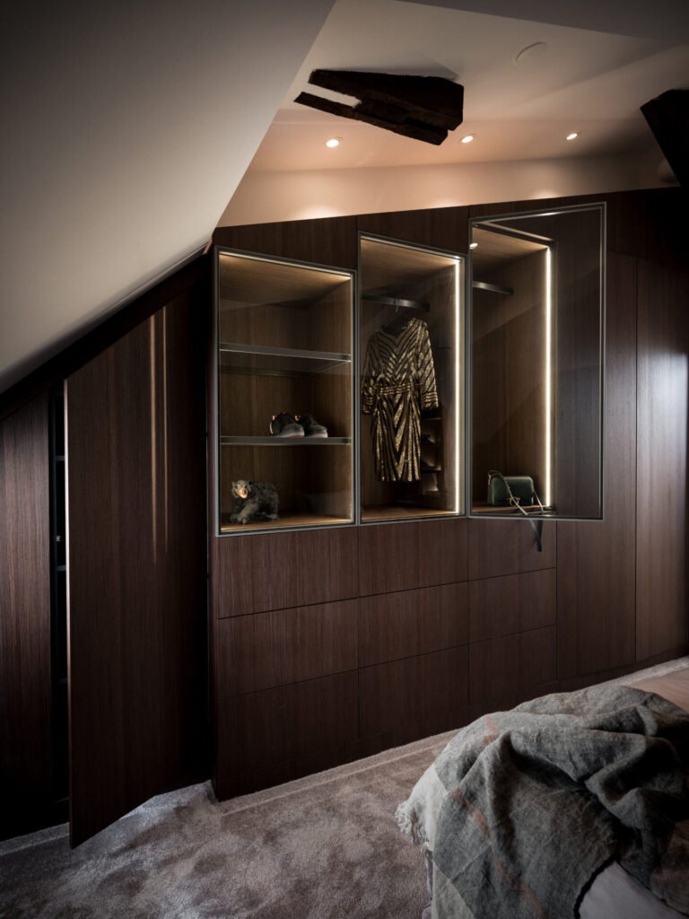 Stiligt - En stiligt trägarderob med glasdisplayer visar upp en klänning, accessoarer och är upplyst av spotlights i ett heltäckningsmatta rum med sluttande tak.