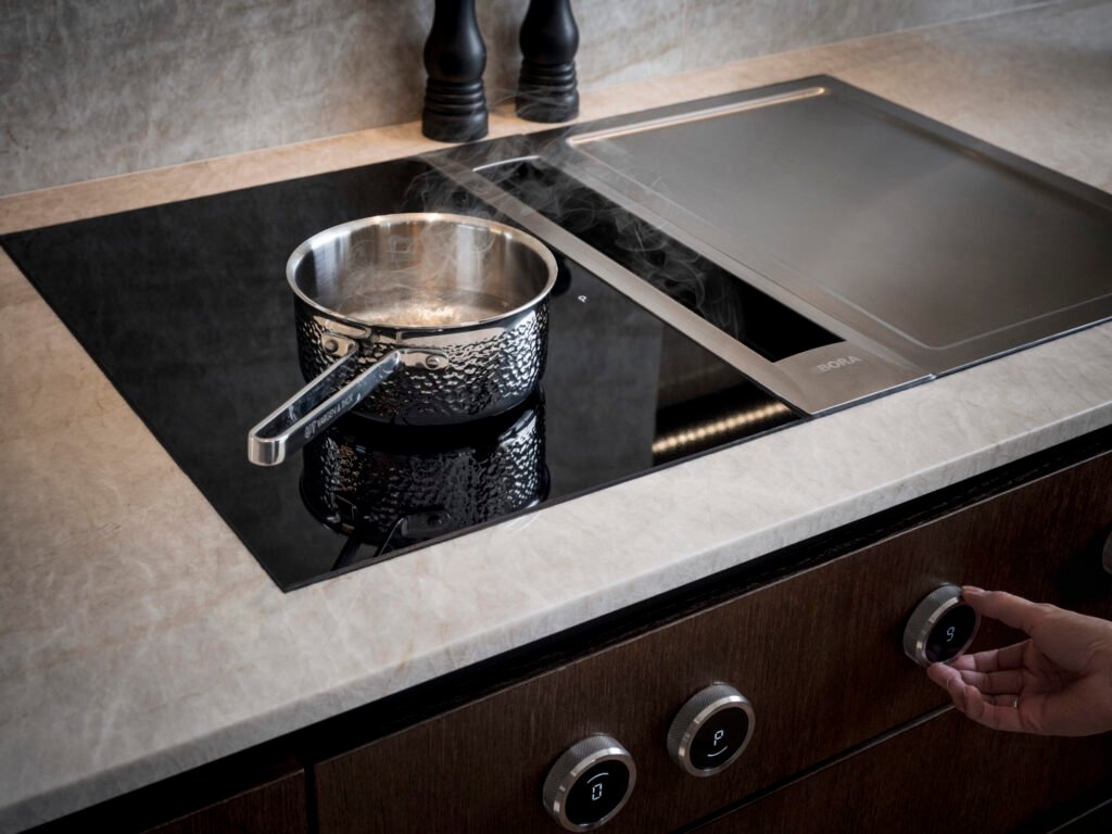 Stiligt - En kastrull med ånga som stiger sitter vackert på en STILIGT induktionshäll. En hand justerar graciöst kontrollvredet, medan två pepparkvarnar står elegant i bakgrunden.