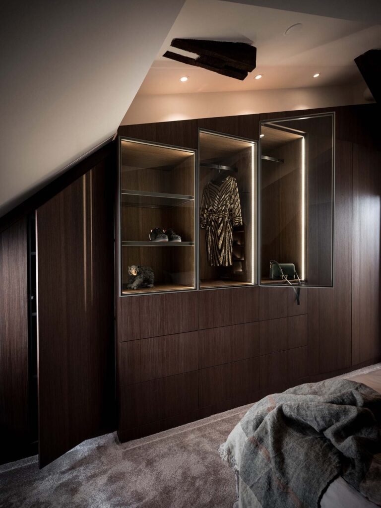 Stiligt - En svagt upplyst garderob med stiligt glasmontrar som visar ett mönstrat plagg, skor och accessoarer. En delvis synlig säng med en grå filt står i förgrunden.