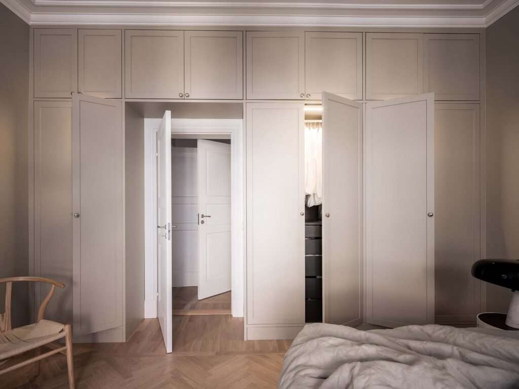 Stiligt - Det stiliga sovrummet har beige inbyggda garderober och öppna dubbeldörrar, med en trästol graciöst placerad till vänster. I förgrunden ger en del av en obäddad säng en avslappnad touch till det eleganta utrymmet.