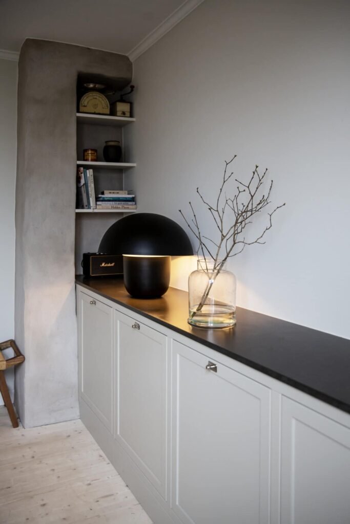 Stiligt - Klassiskt shakerkök en stiligt svart bänkskiva, dekorativa grenar i en glasvas, böcker på vita hyllor och mjuk belysning.