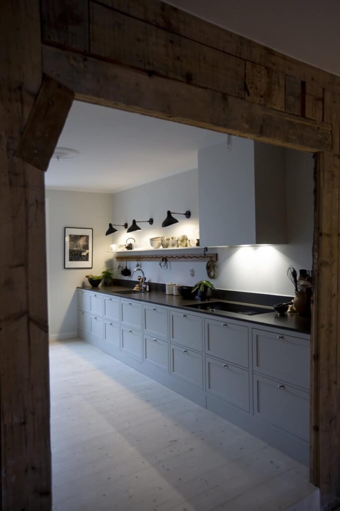 Stiligt - Ett modernt stiligt kök har en lång bänkskiva, belysning under skåpet och tre snygga svarta vägglampor. Trädekor och hyllor fulla av olika köksartiklar kompletterar den chica atmosfären.