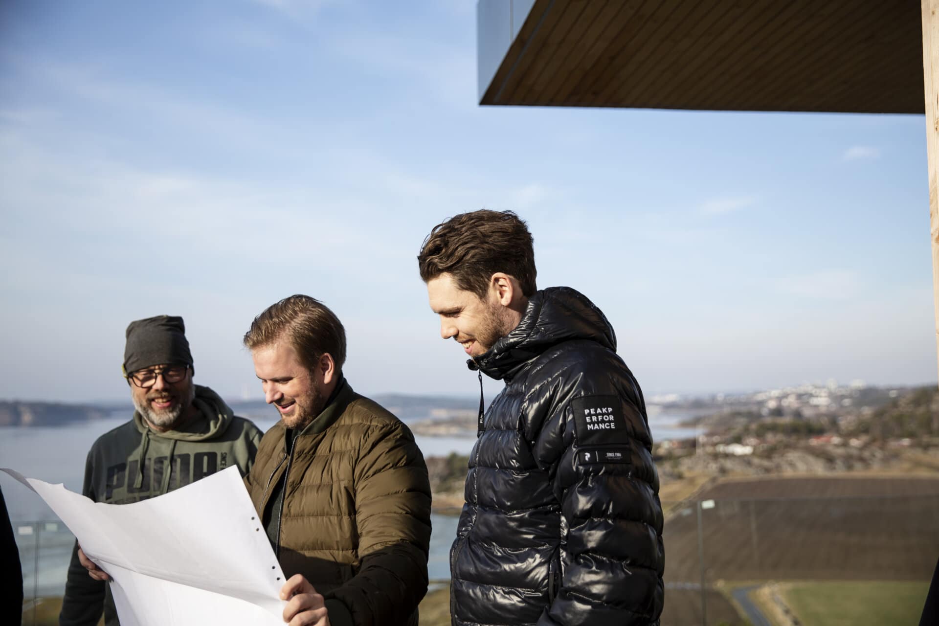 Stiligt - Tre män i stiliga vinterjackor undersöker arkitektoniska planer utomhus, inramade av en naturskön bakgrund.