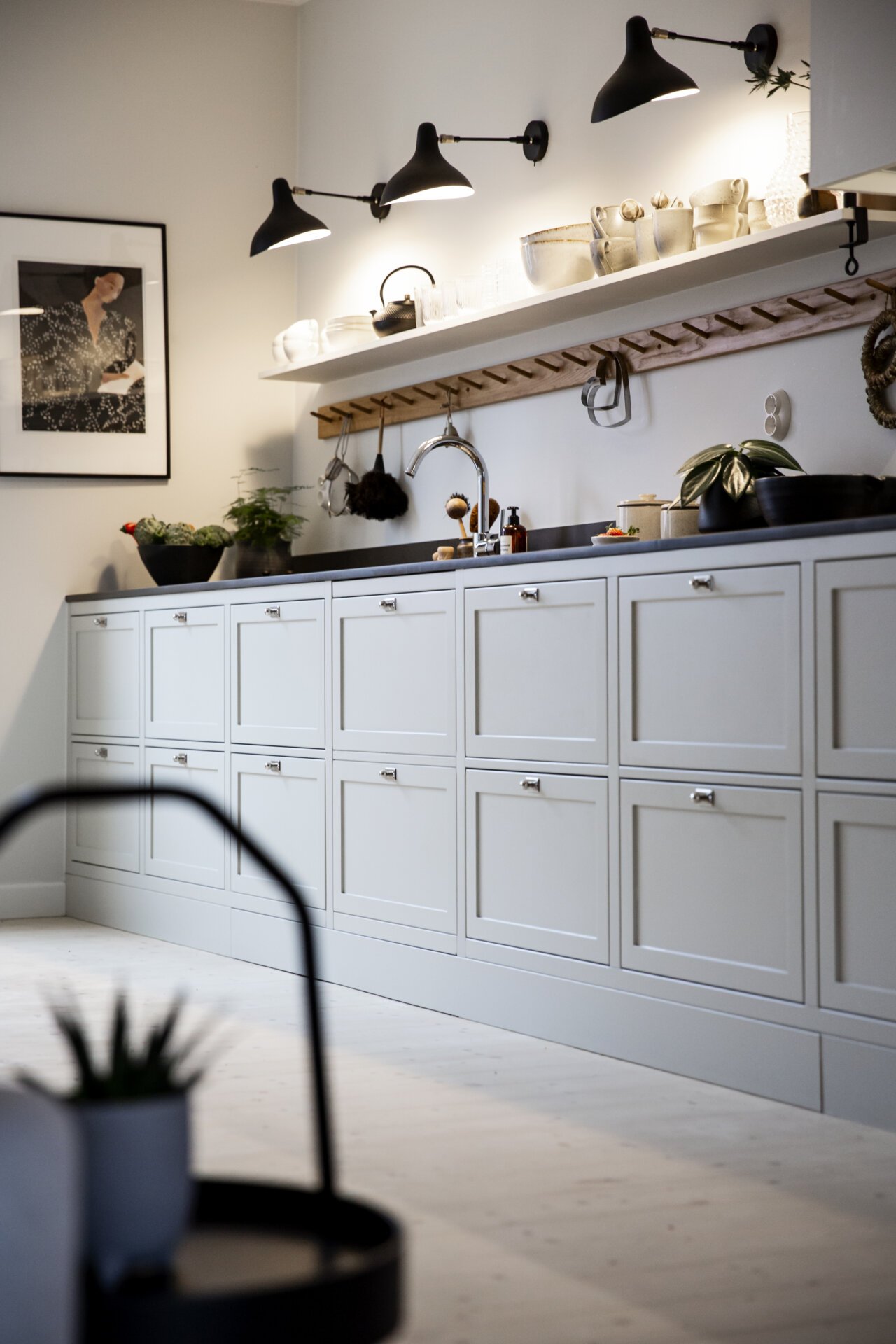 Stiligt - Ett modernt kök ståtar med stiligt ljusgrå skåp, öppna hyllor fyllda med porslin och väggmonterade svarta lampor. En inramad bild pryder väggen medan en suddig krukväxt ger en touch av natur i förgrunden.