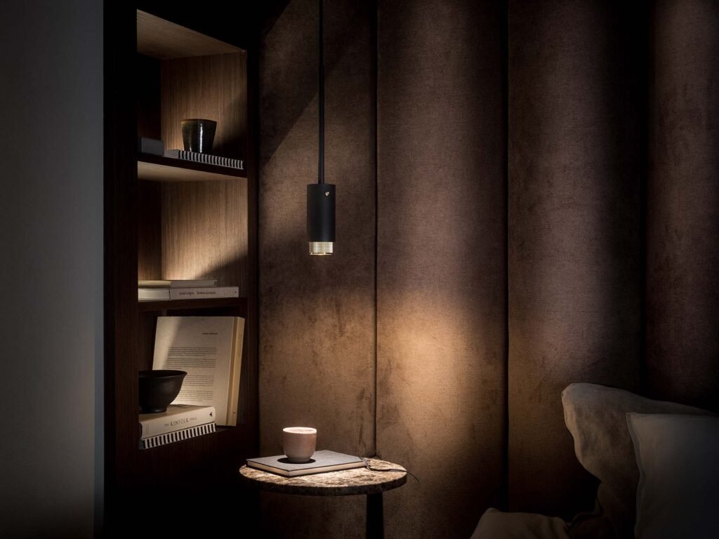 Stiligt - Ett mysigt rum med en stiligt upplyst pendel, ett litet bord med en kopp och en bokhylla prydd med böcker och dekor. Miljön är mjukt upplyst, vilket skapar en varm atmosfär.