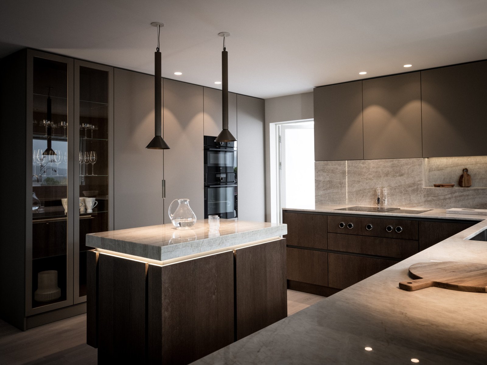 Stiligt - Modernt kök med varma färger, mörka träskåp, marmorbänkskiva på köksön, integrerade vitvaror, eleganta pendellampor och glas i vitrinskåp.