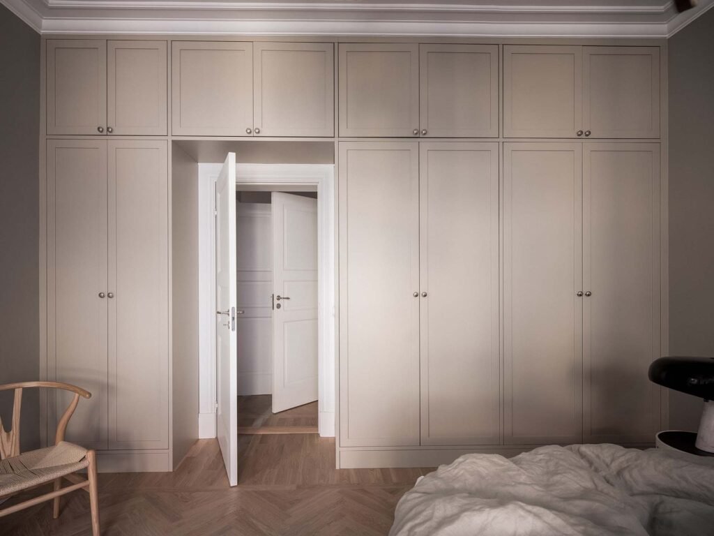 Stiligt - Stängda inbyggda garderober kantar en sovrumsvägg med en dörr på glänt och utstrålar en stiligt charm. En säng med fräscha vita sängkläder och en elegant enkel trästol kompletterar denna sofistikerade miljö.