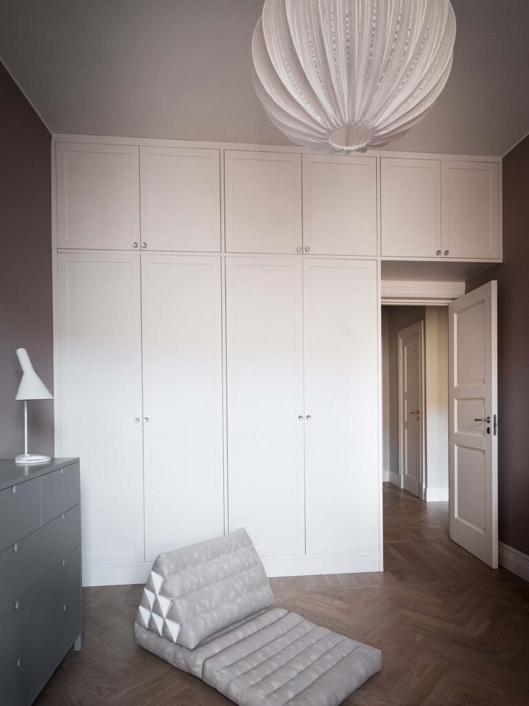 Stiligt - Det stiligt minimalistiska rummet har stora vita garderober, en hög grå byrå och en modern triangulär golvkudde. En vit lampa kompletterar utrymmet, medan en sfärisk taklampa hänger ovanför. Dörren står lite på glänt.