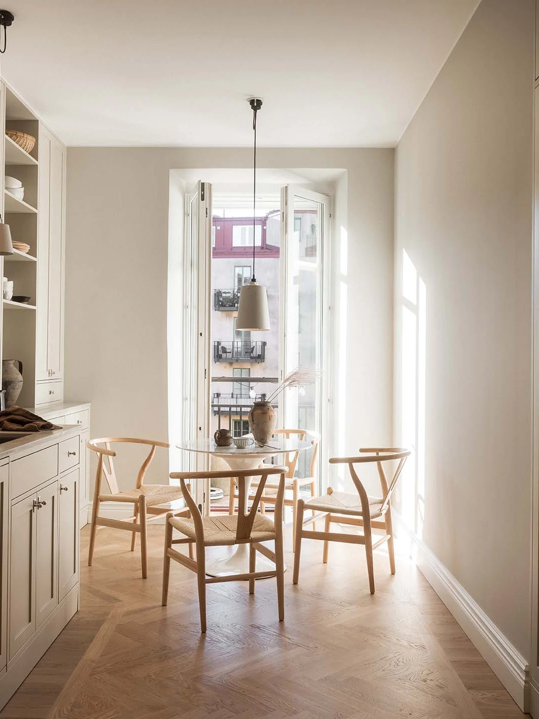 Stiligt - En minimalistisk matplats med ett runt glasbord, fyra trästolar i klassisk Shaker-stil, en taklampa och naturligt ljus som strömmar in genom höga, öppna fönster med utsikt över flerbostadshus.