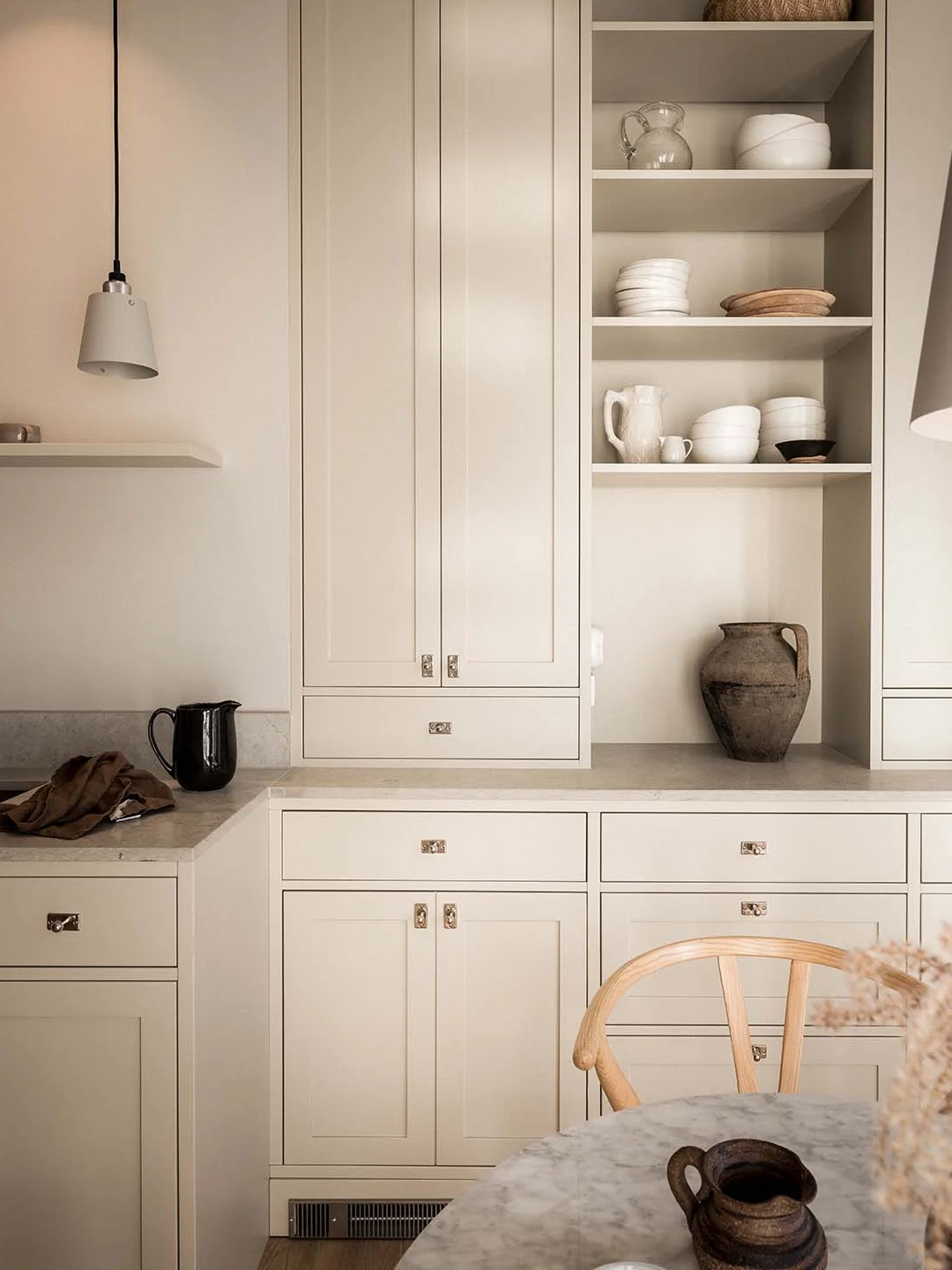 Stiligt - Ett minimalistiskt beige kök i Shakerstil med öppna hyllor där vita tallrikar och keramik står framme, stängda skåp nedanför, bänkskiva i marmor, en svart kanna och en trästol i förgrunden.