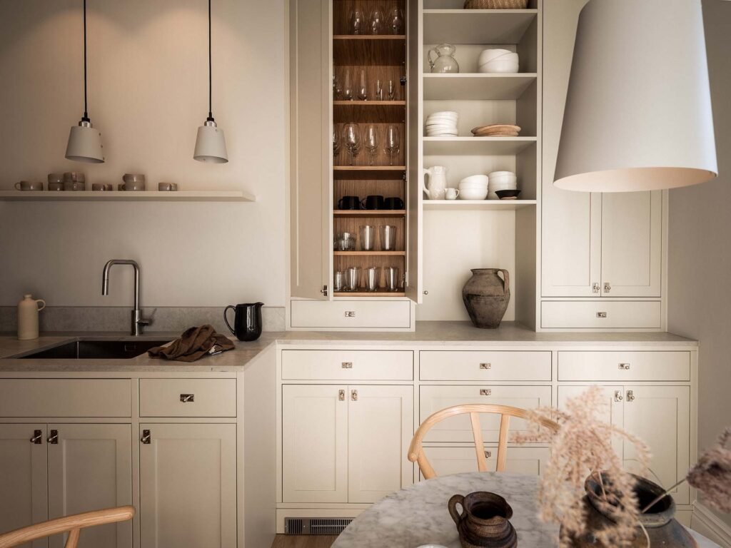 Stiligt - Ett stiligt modernt kök har beiga skåp och öppna hyllor som visar upp rätter, kompletterat med eleganta armaturer. I förgrunden fullbordar ett runt bord prydt med torkade blommor och keramiska föremål detta eleganta utrymme.