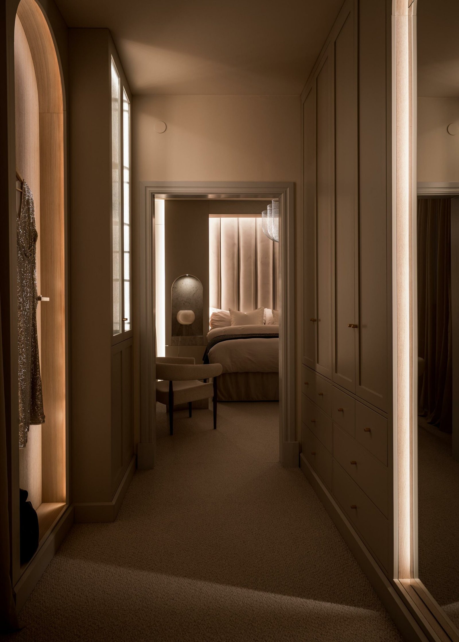Stiligt - Ett svagt upplyst sovrum med stiliga neutrala toner visar upp en säng med vadderad sänggavel, en elegant stol och inbyggda garderober. En klänning hänger elegant i en nisch, badad i mjuk belysning.