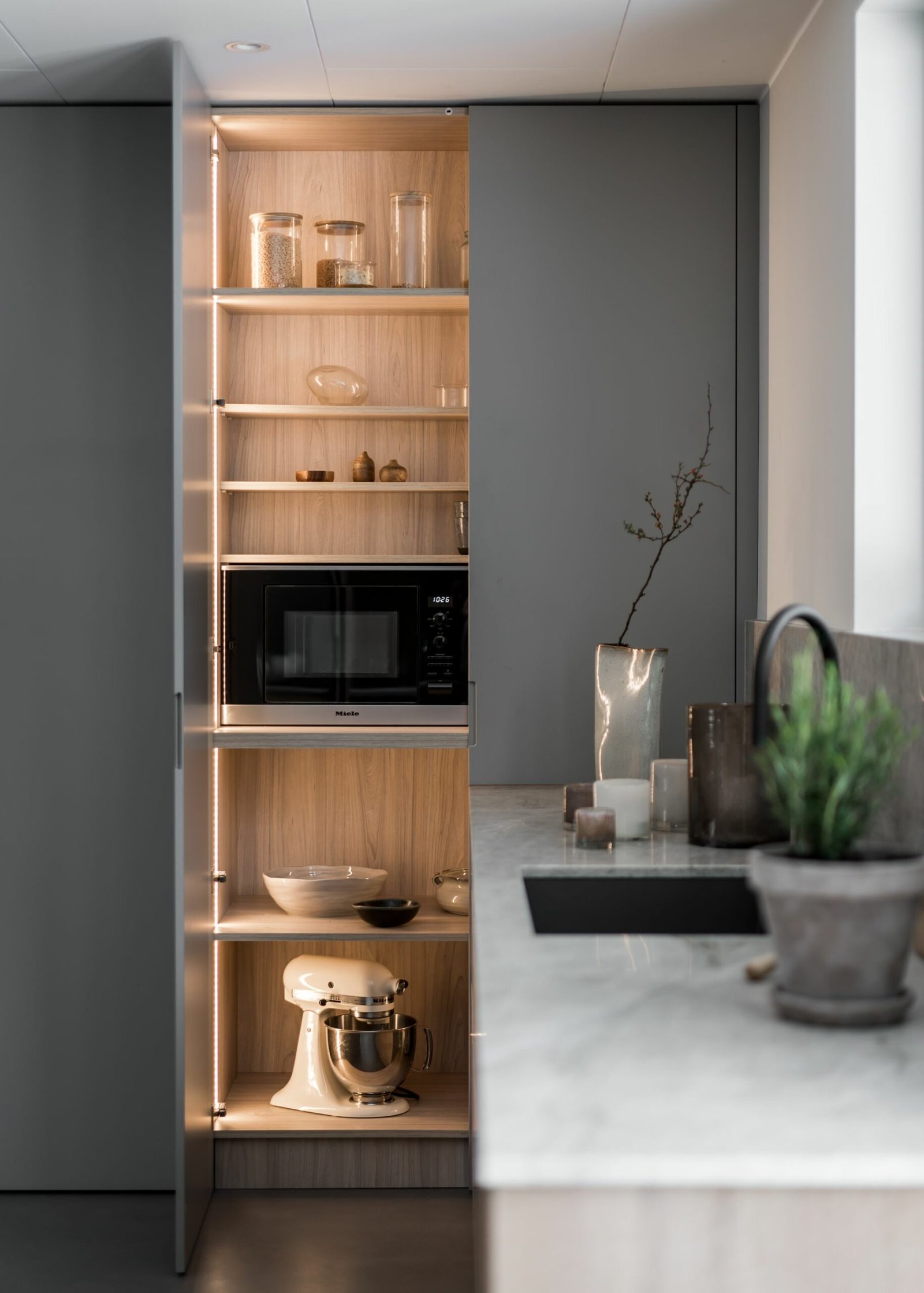 Stiligt - Ett stiligt köksskafferi med öppna hyllor visar upp en mikrovågsugn, ställblandare, glasburkar och dekorativa föremål. De bakgrundsbelysta hyllorna lyser upp utrymmet vackert, medan bänkskivan är prydd med en växt och diverse köksartiklar.