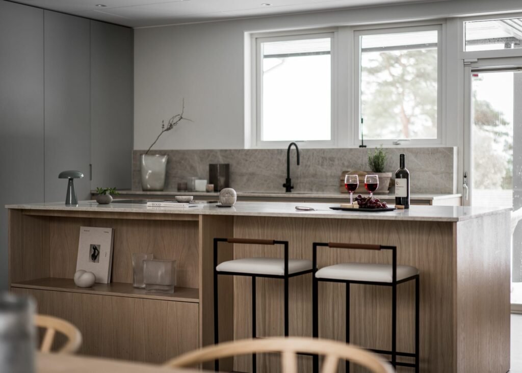 Stiligt - Ett modernt stiligt kök har en ö i ljust trä tillsammans med två eleganta pallar och smakfulla prydnadsföremål. En flaska vin och två glas sitter lockande på bänken medan solljuset mjukt silar in genom fönstren.