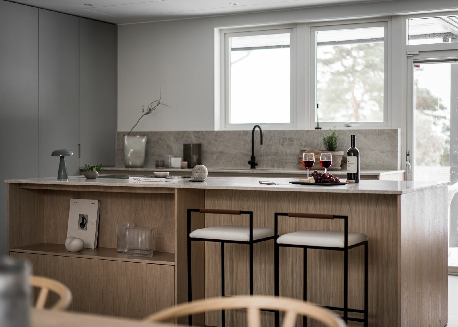 Stiligt - Ett modernt stiligt kök har en ö i ljust trä tillsammans med två eleganta pallar och smakfulla prydnadsföremål. En flaska vin och två glas sitter lockande på bänken medan solljuset mjukt silar in genom fönstren.