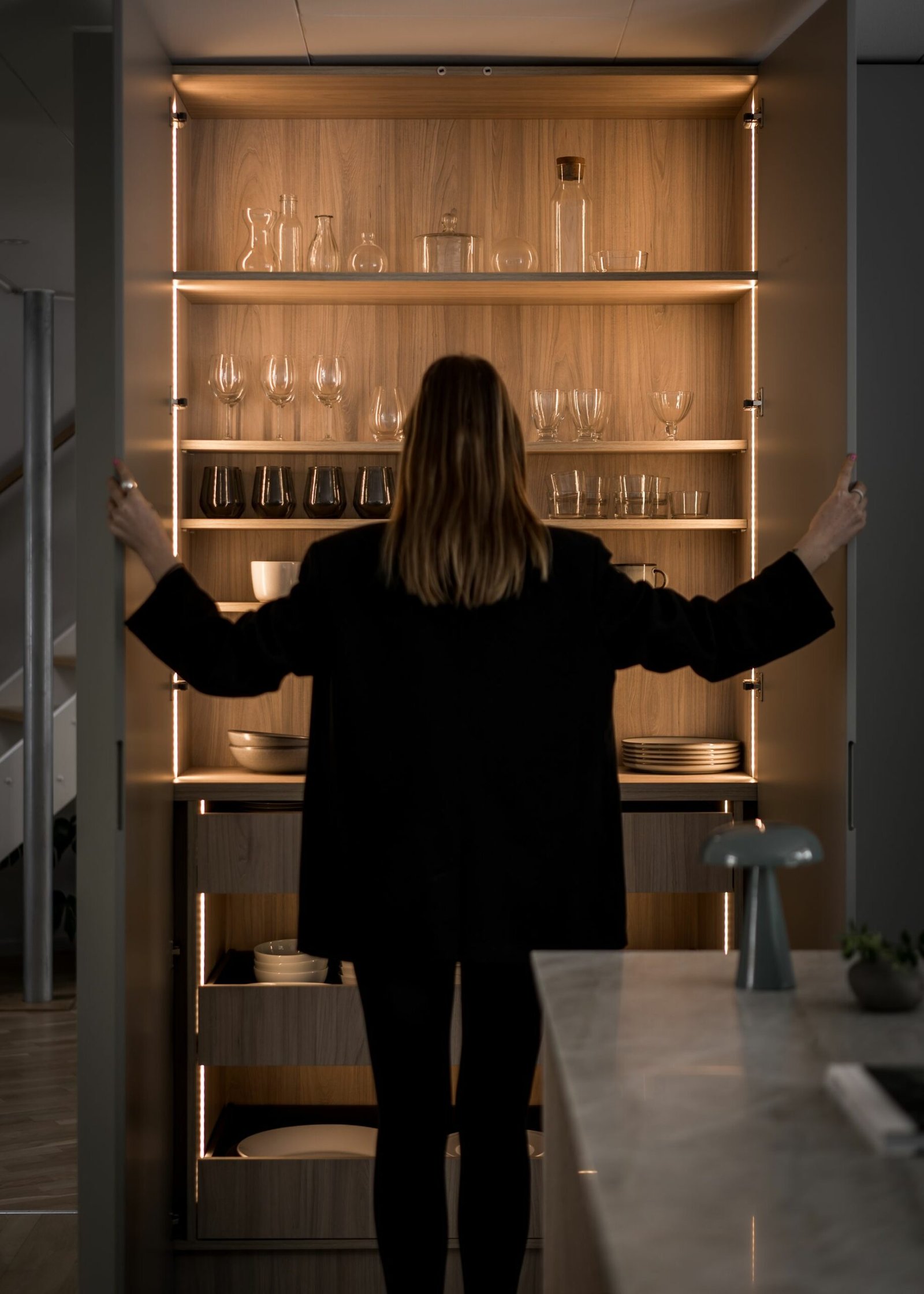 Stiligt - I en stiligt modern köksmiljö öppnar en person i mörka kläder ett bakgrundsbelyst skåp fyllt med eleganta glasföremål och eleganta fat.