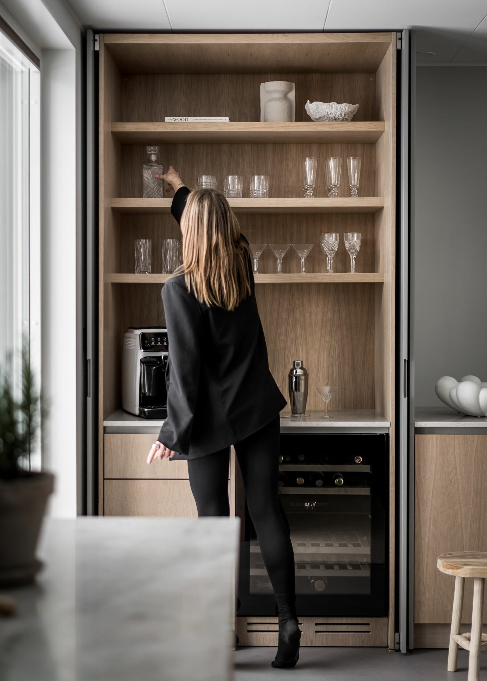 Stiligt - En kvinna i stiligt svart outfit sträcker sig efter ett glas på en kökshylla i trä, omgiven av en kaffemaskin, eleganta glasföremål och en elegant vinkyl.