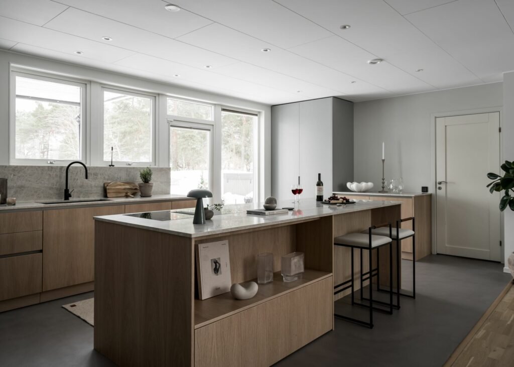 Stylish - Modern kitchen with wooden cabinets and a stylish island counter along with sleek stools, wine glasses and tasteful decor items. Large windows flood the space with natural light, while a door provides easy outdoor access. The design remains minimalist yet stylishly functional.