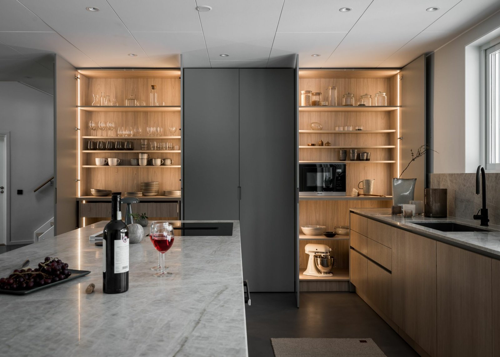 Stiligt - Ett stiligt modernt kök har bänkskivor i marmor och öppna hyllor som visar eleganta glas och porslin, med ett fönster till höger. En flaska vin och ett glas står inbjudande på bänken.