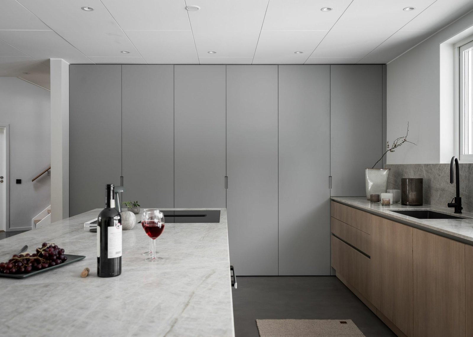 Stiligt - Ett modernt kök har en marmorö prydd med ett glas rött vin, en flaska och en tallrik med druvor. Stiligt gråa skåp och underskåp i trä kantar väggarna medan ett fönster ovanför diskbänken bjuder in naturligt ljus att flöda in.