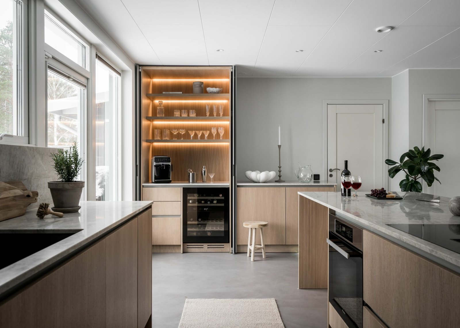 Stiligt - Stiligt modernt kök med ljusa träskåp, glas på upplysta hyllor, en espressomaskin, och en vinflaska med glas på bänken. Stora fönster flödar över utrymmet med naturligt ljus.