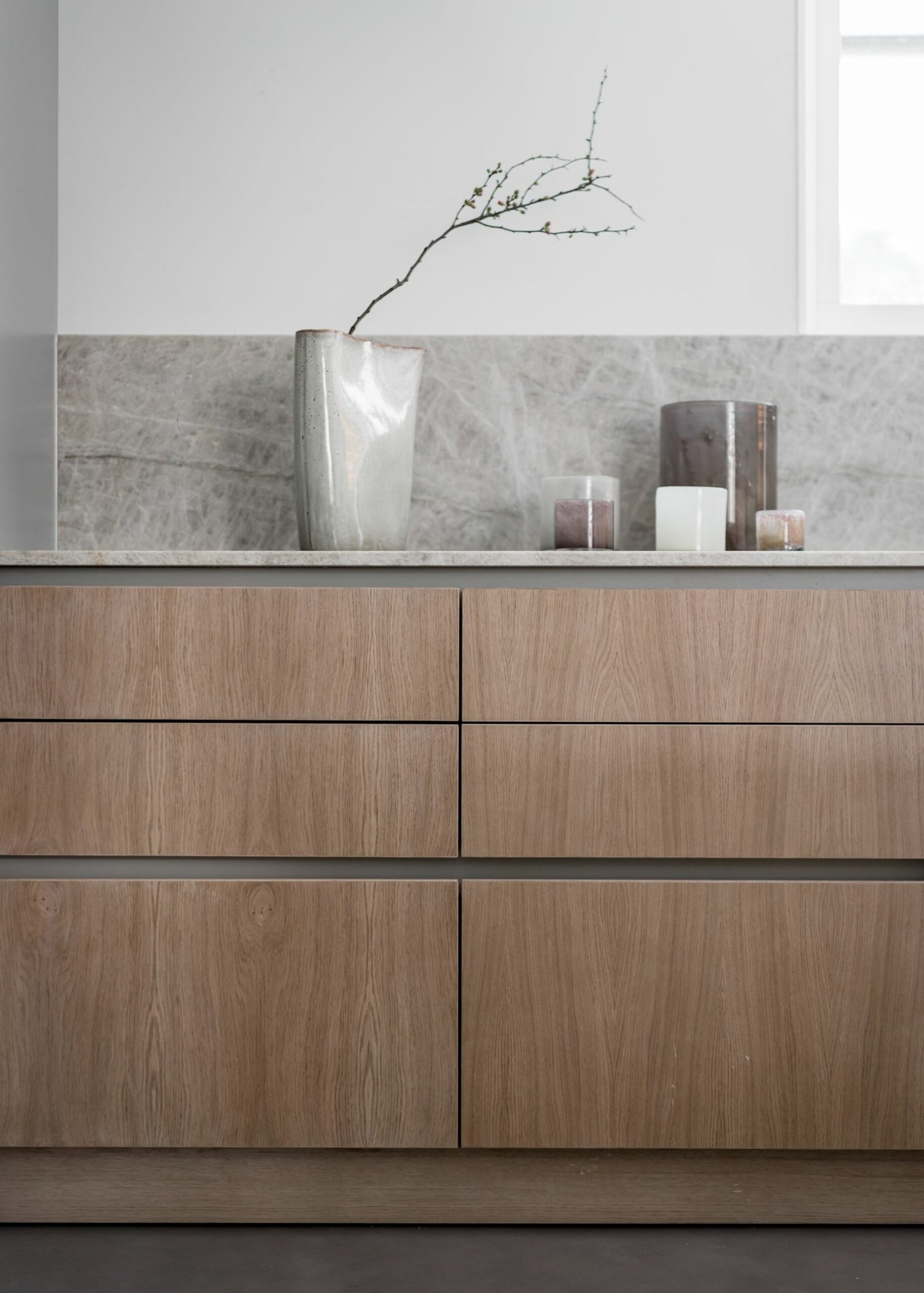 Stiligt - En stiligt minimalistisk köksbänk har skåp i ljust trä, en hög vas med grenar och flera ljus inställda mot en grå marmorbaksida.