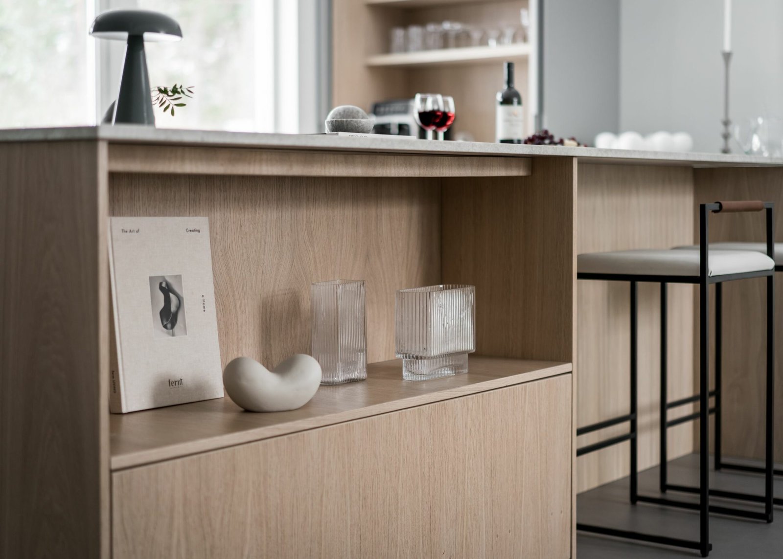 Stiligt - Ett stiligt minimalistiskt kök med trähyllor visar dekorativa föremål, en flaska vin och ett glas på bänkskivan. Moderna barstolar är strategiskt placerade i närheten, vilket förstärker dess eleganta estetik.