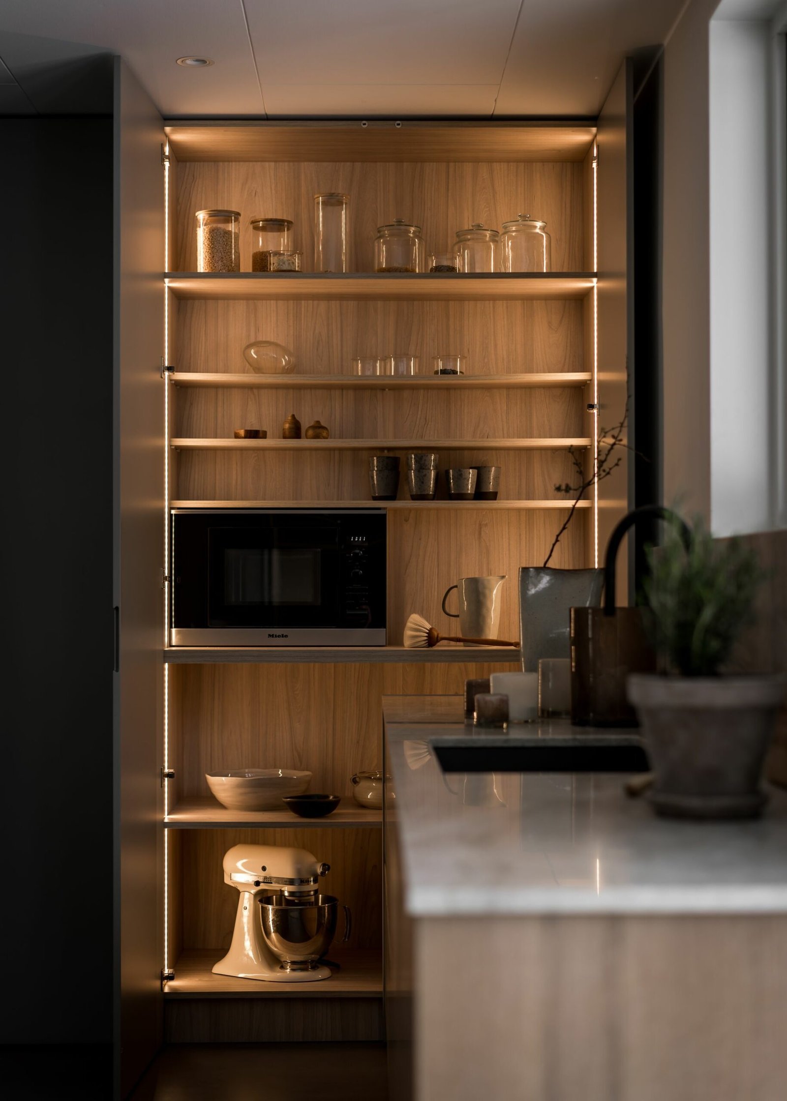 Stiligt - Kökshyllor i trä, arrangerade med stilistisk stil, displayburkar, mikrovågsugn, koppar och mixer. Mjuk belysning framhäver stilleben.