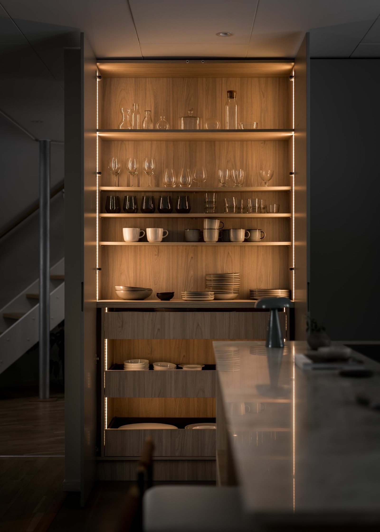 Stiligt - Ett svagt upplyst, stiligt träskåp med glashyllor visar en elegant samling av glasvaror, koppar och tallrikar i en modern köksmiljö.