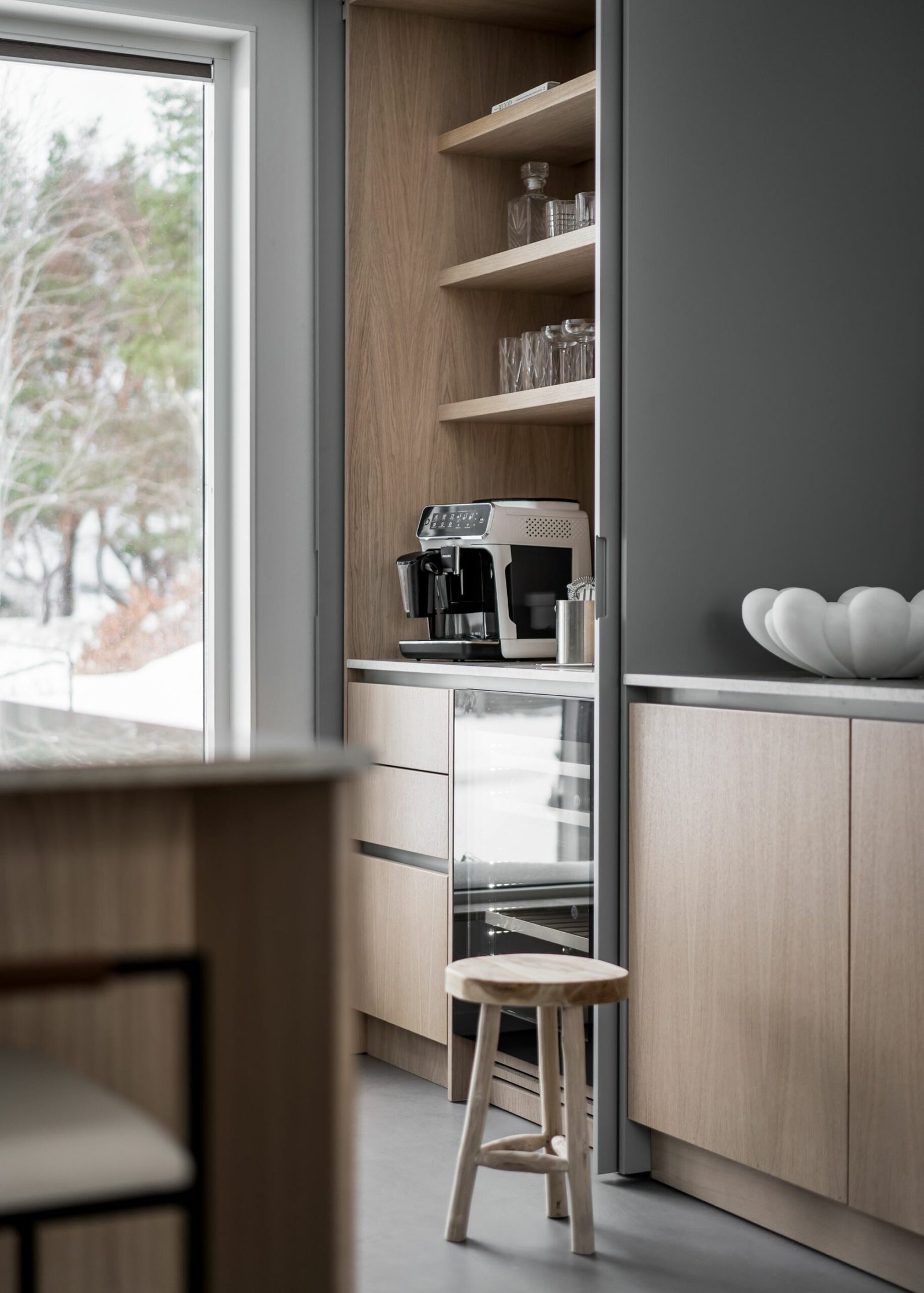 Stiligt - Modernt kök med stiligt ljusa träskåp, en inbyggd kaffemaskin, glasföremål på hyllor, en liten träpall och ett fönster som visar upp en snöig utomhusvy.