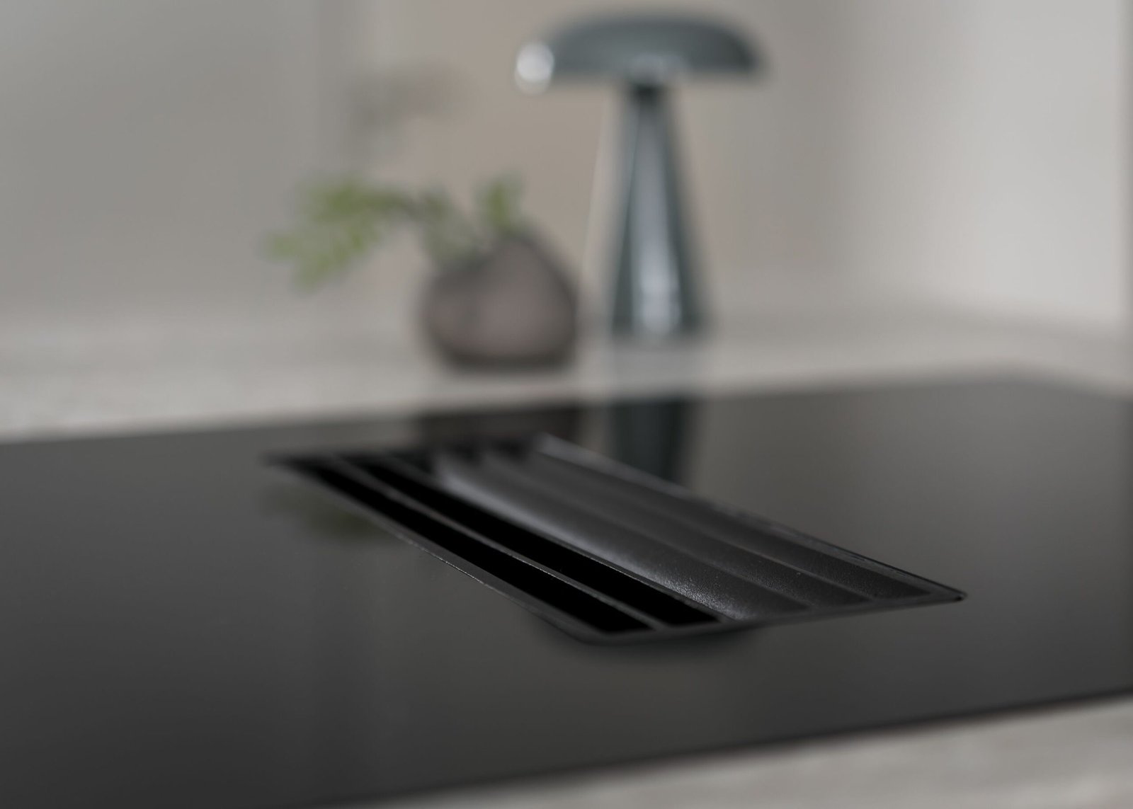 Stiligt - Närbild av en elegant, stilig spishäll med en central ventilationspanel. I den suddiga bakgrunden bidrar en växt och lampa till den moderna atmosfären.