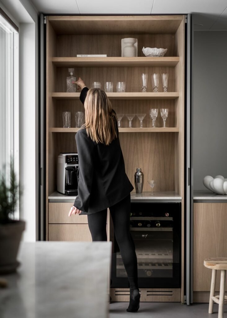 Stiligt - En kvinna i stiligt svart outfit sträcker sig efter ett glas på en kökshylla i trä, omgiven av en kaffemaskin, eleganta glasföremål och en elegant vinkyl.