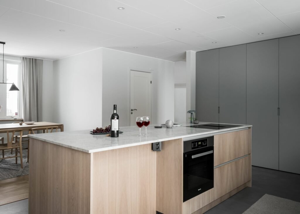 Stiligt - Modernt kök med en ljus träö, bänkskiva i marmor, induktionsspis och inbyggd ugn. En flaska vin, två glas och vindruvor pryder disken elegant. Den stiliga matplatsen syns inbjudande i bakgrunden.