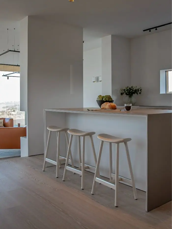 Stiligt - Fernströms minimalistiska kök präglas av en snövit köksö med tre matchande barstolar, en fruktskål och en liten vas med blommor. Den öppna planlösningen kopplar smidigt samman köket med ett elegant vardagsrum i bakgrunden.