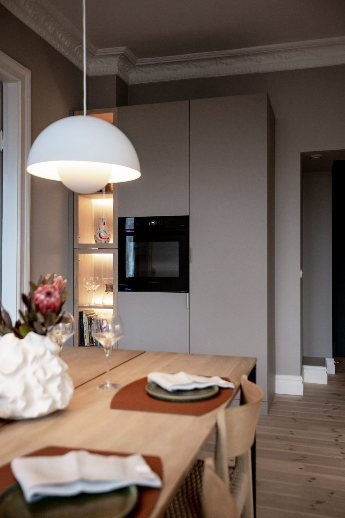 Stiligt - En modern matplats med ett träbord dukat för två, en taklampa ovanför och inbyggda köksskåp med ugn och upplysta hyllor i bakgrunden – ett elegant modernt kök i en sekelskifteslägenhet som på ett perfekt sätt förenar klassisk charm med modern design.
