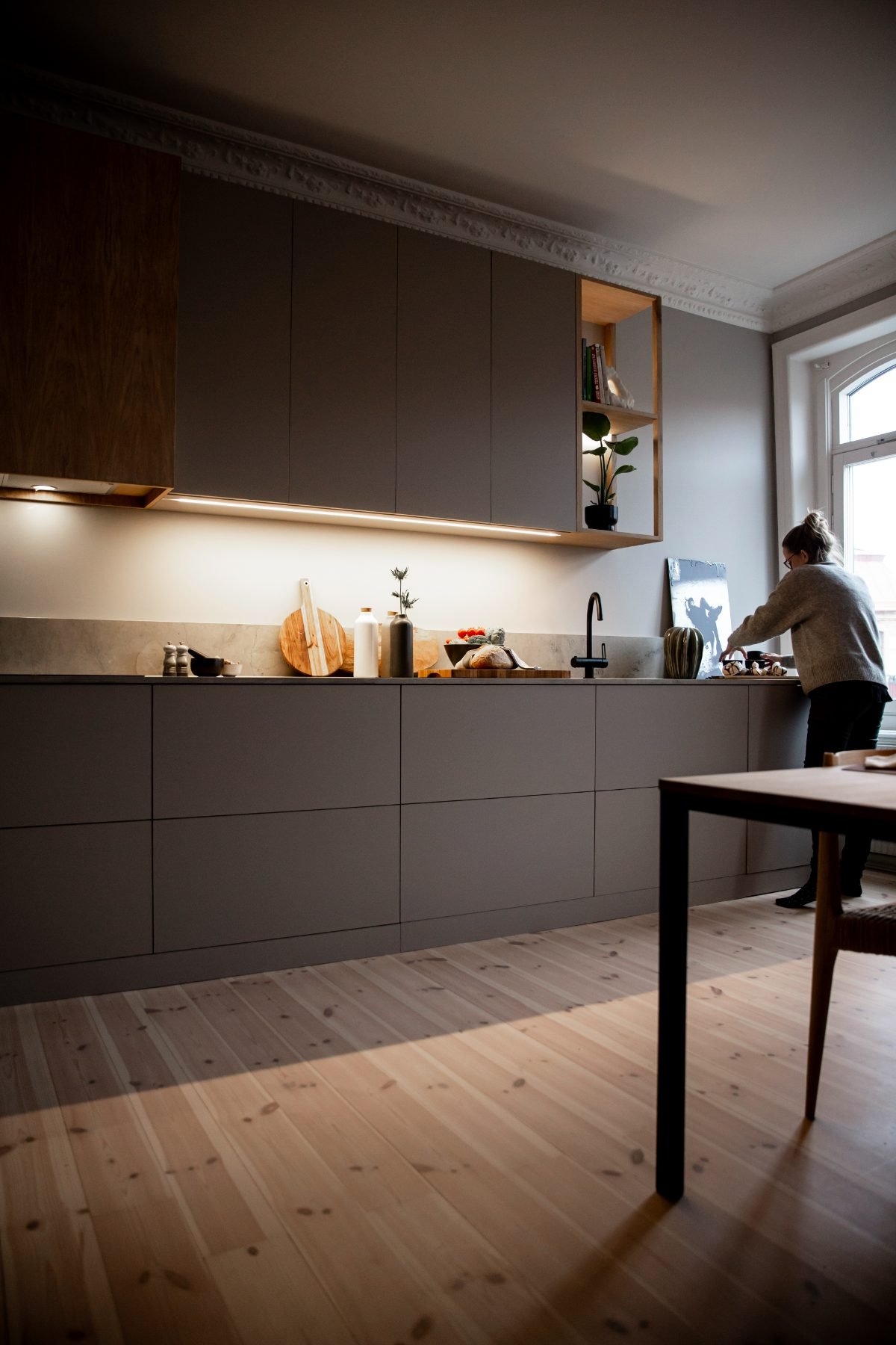 Stiligt - En person står vid ett diskbänk i ett modernt kök med gråa skåp, inslag av trä och ett stort fönster som släpper in naturligt ljus, vilket är ett exempel på ett modernt kök i en sekelskifteslägenhet.