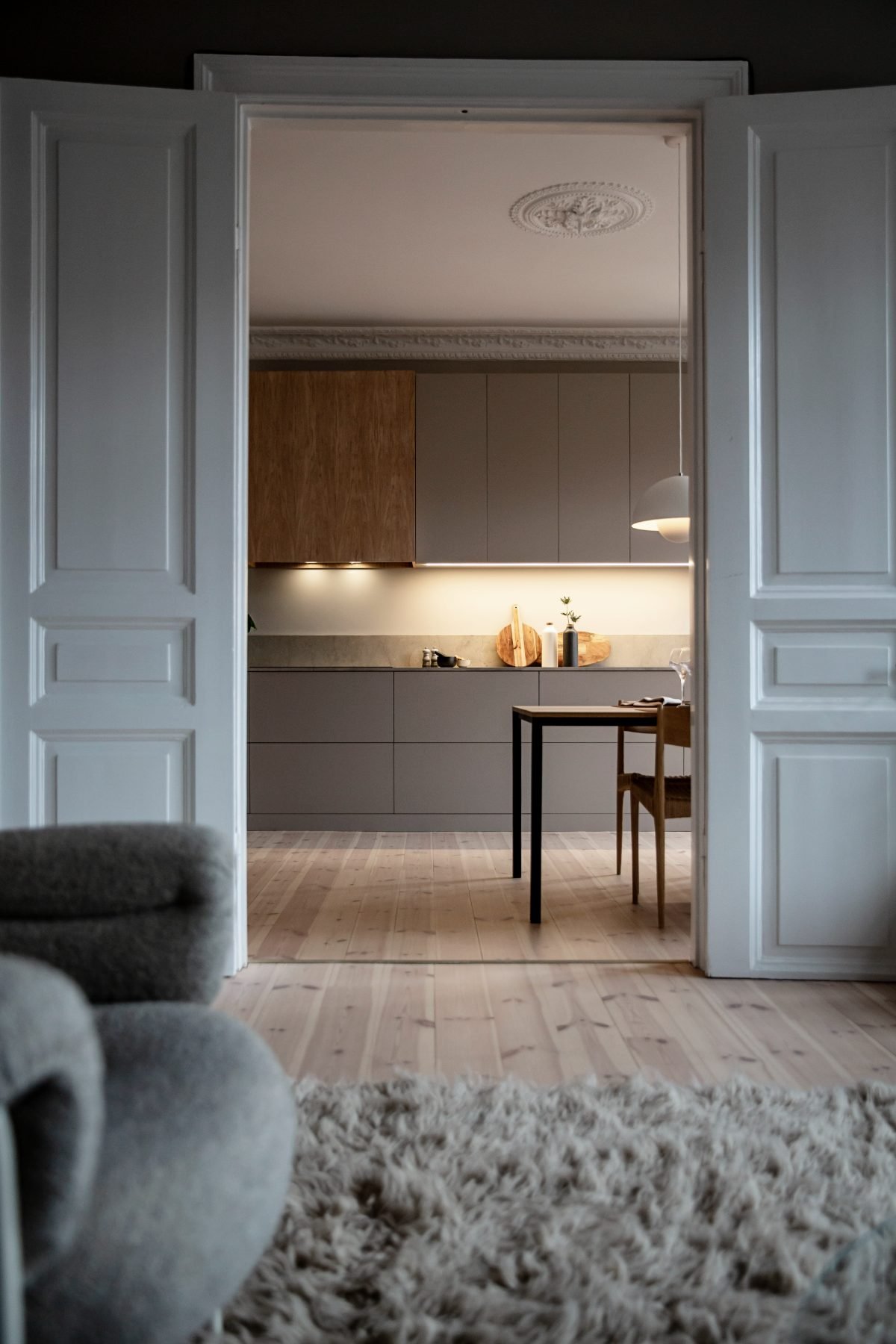 Stiligt - Utsikt genom öppna dubbeldörrar in i ett modernt kök i en sekelskifteslägenhet med gråa skåp, trädetaljer, ett matbord och en taklampa; en mjuk matta och en fåtölj i förgrunden.