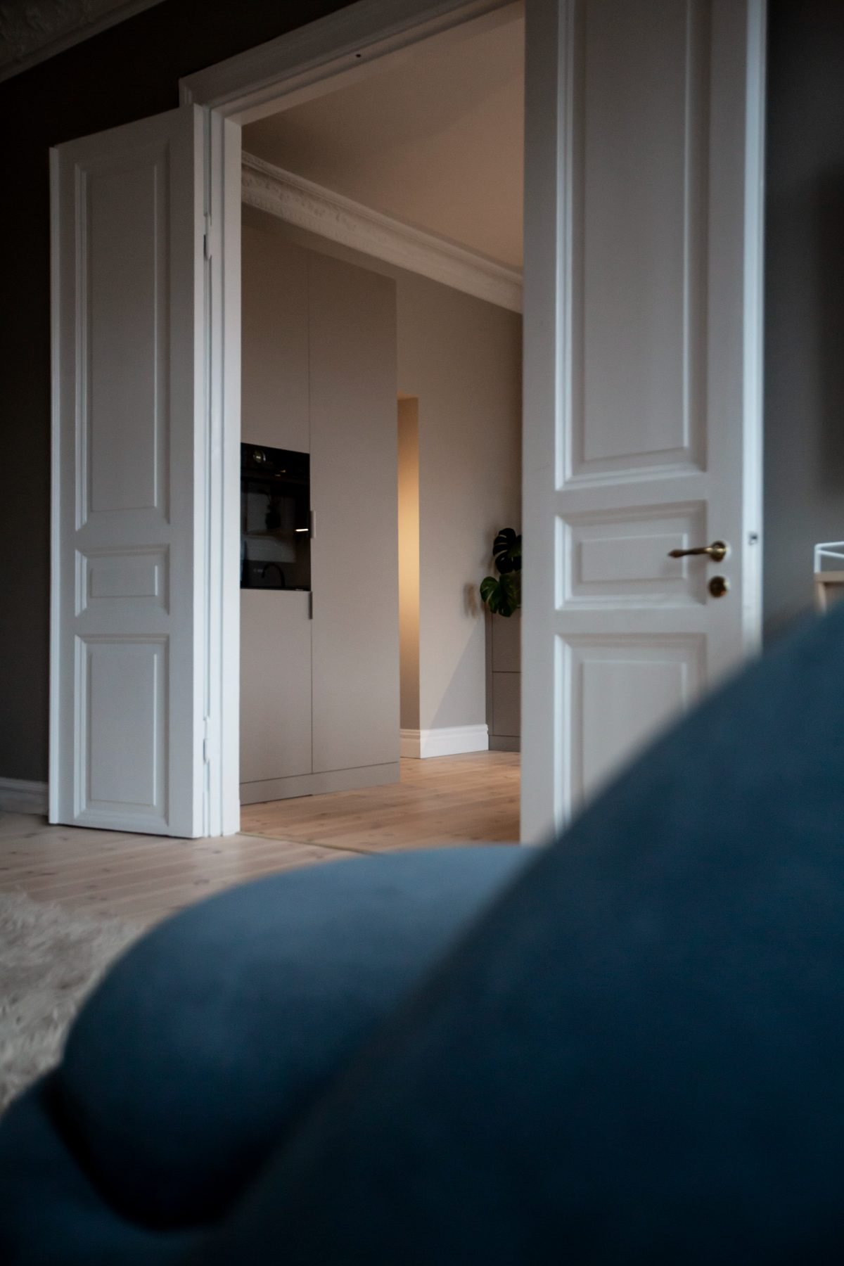 Stiligt - När man öppnar de vita dubbeldörrarna möts man av en hall med ljusa trägolv, neutrala väggar och en delvis utsikt över ett modernt kök i en sekelskifteslägenhet, med eleganta köksskåp och en växt i bakgrunden.