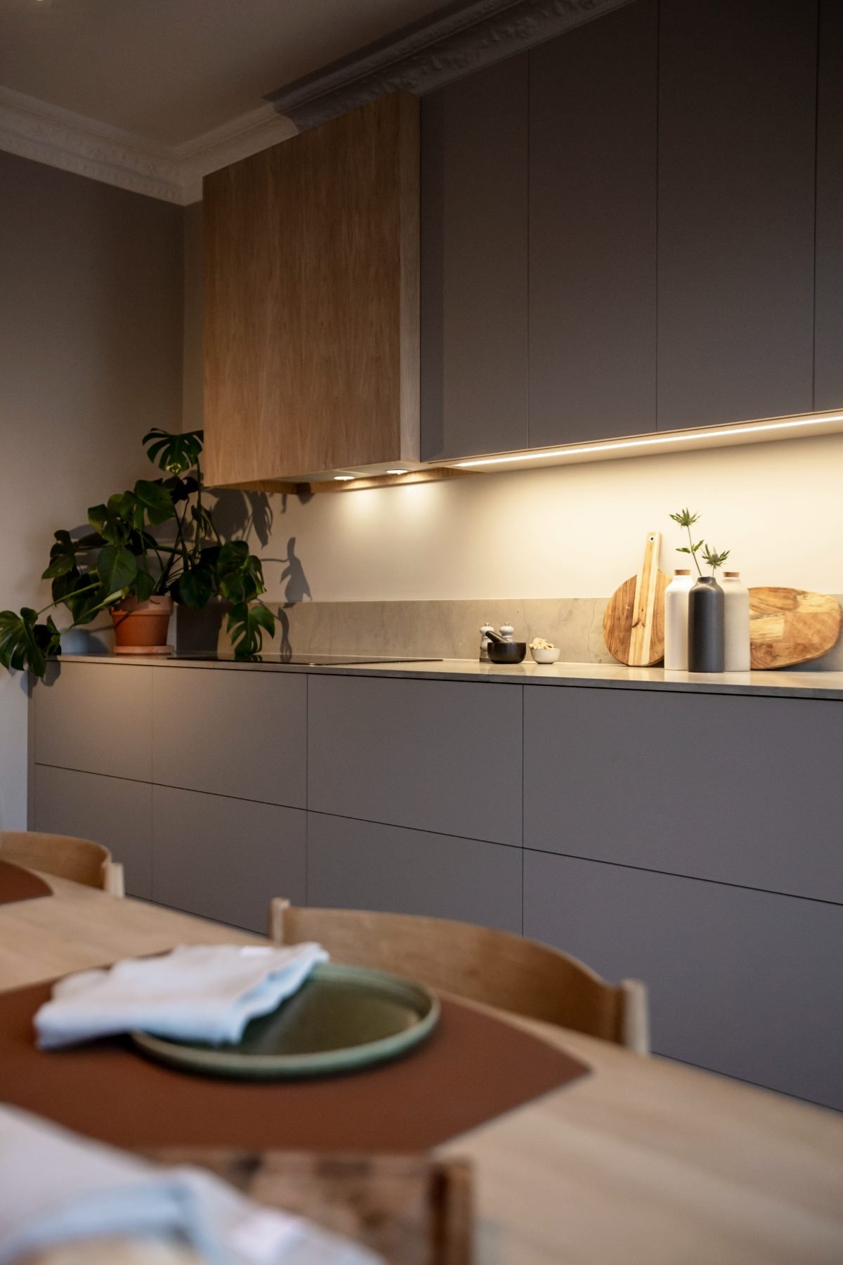 Stiligt - Ett modernt kök med gråa skåp, detaljer i trä, en krukväxt, skärbrädor och ett snyggt dukat matbord med tallrikar och servetter. Den varma belysningen under skåpen förstärker den inbjudande stämningen i detta moderna kök i en sekelskifteslägenhet.