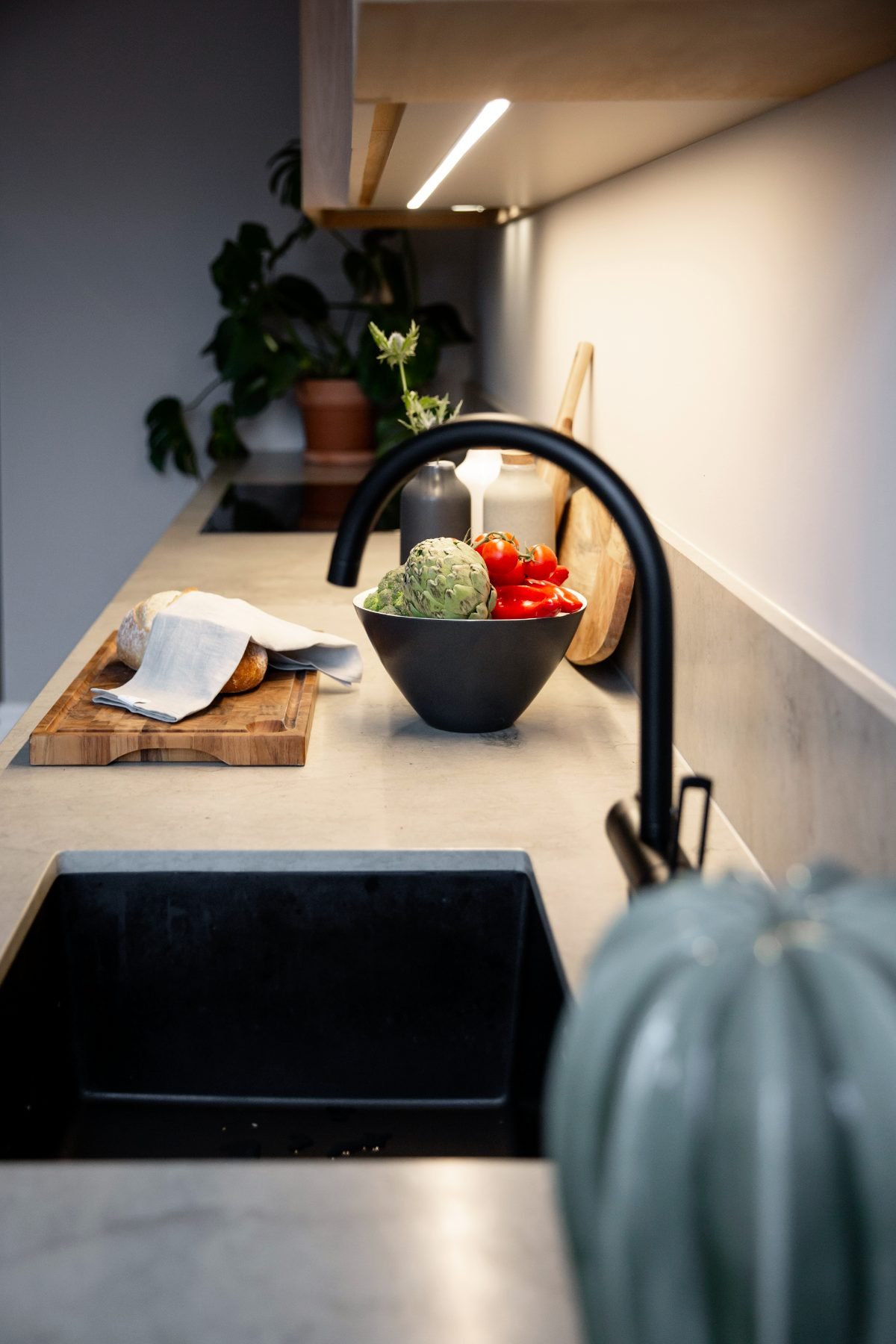 Stiligt - En modern köksbänk med en svart diskho, en skål med grönsaker, en skärbräda med bröd och krukväxter i bakgrunden – en perfekt blandning av modern bekvämlighet och tidlös charm i ett modernt kök i en sekelskifteslägenhet.