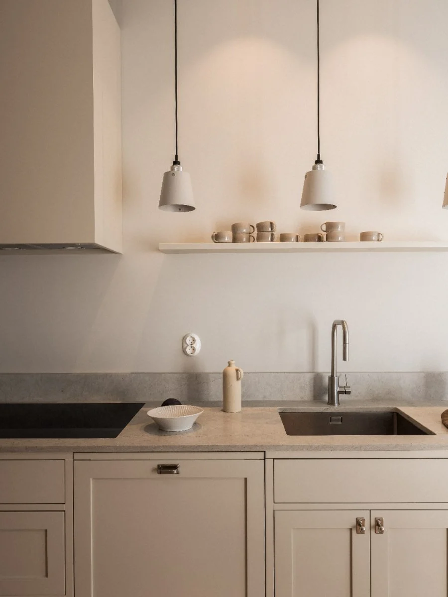 Stiligt - Ett minimalistiskt kök i Shakerstil med beige skåp, grå bänkskivor, två diskhoar, tre taklampor och en hylla med koppar och skålar mot en vit vägg.