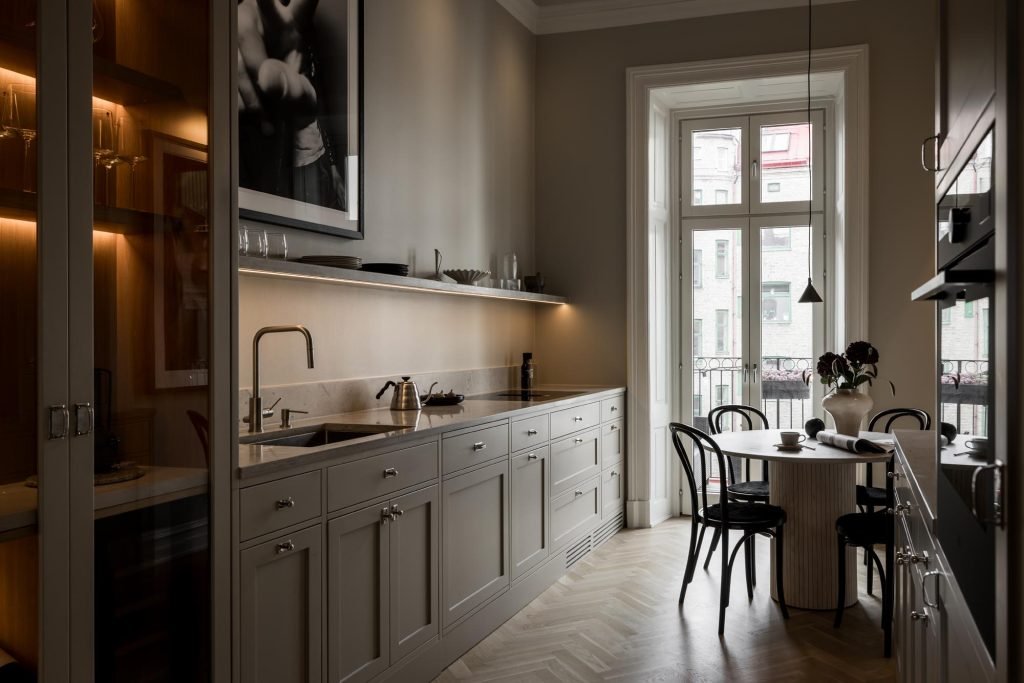 Stiligt - Ett modernt stiligt kök med ljusa skåp, en inbyggd ugn och ett runt matbord tillsammans med svarta stolar. Ett stort fönster välkomnar naturligt ljus medan konst dekorerar väggen elegant.