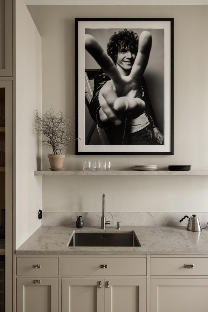 Stiligt - Stiligt köksinredning med bänkskiva i marmor, diskbänk och vattenkokare. Ett svartvitt foto av en person som gör ett fredstecken är inramat på väggen ovanför en hylla prydd med glasögon och en krukväxt.