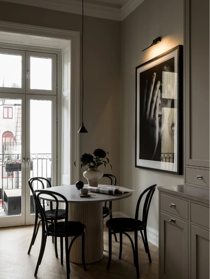 Stiligt - En liten matplats med ett runt bord, fyra svarta stolar, en taklampa, ett stort fotografi av en hand och ett fönster med utsikt över balkongen smälter sömlöst samman med ett kök i shakerstil och skapar en inbjudande och elegant atmosfär.