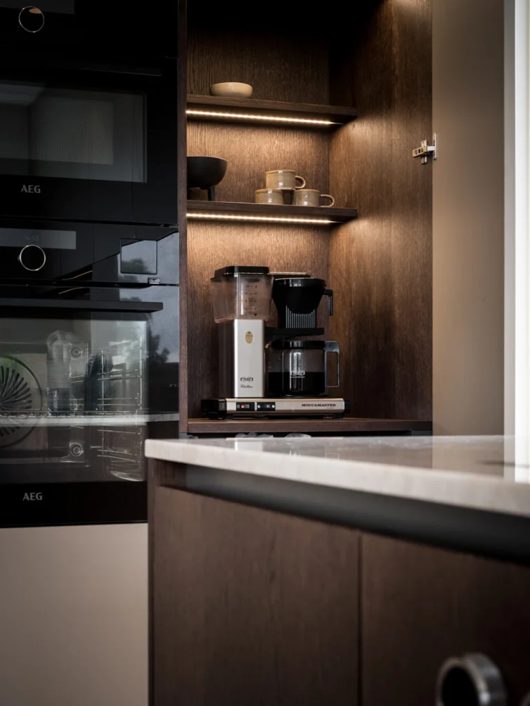 Stiligt - I ett modernt kök med mörka träskåp och inredning varma färger står kaffebryggare och koppar på öppna hyllor och glasen är synliga bakom en skåpslucka. Inredning Varma Färger: Skapa Hemmets Mysiga Atmosfär