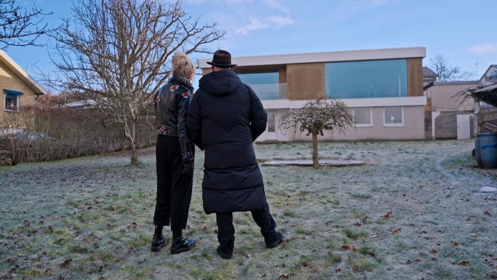 Stiligt - Två personer står på en frostig gräsmatta framför ett modernt hus med stora fönster, som i Husdrömmar 2026 Säsong 13 avsnitt 3, omgivet av kala träd under en klar himmel.