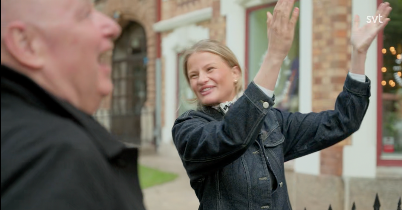 Stiligt - En kvinna i jeansjacka gestikulerar med uppsträckta händer medan hon ler och pratar med en man utanför en tegelbyggnad.